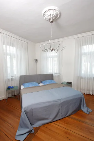 Zimmer mit Doppelbett, zwei hohen Fenstern und Stuckrosette an der Decke