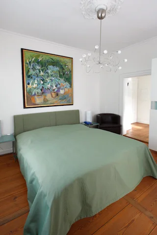 Schlafzimmer mit grüner Tagesdecke und Blumenbild über dem Bett